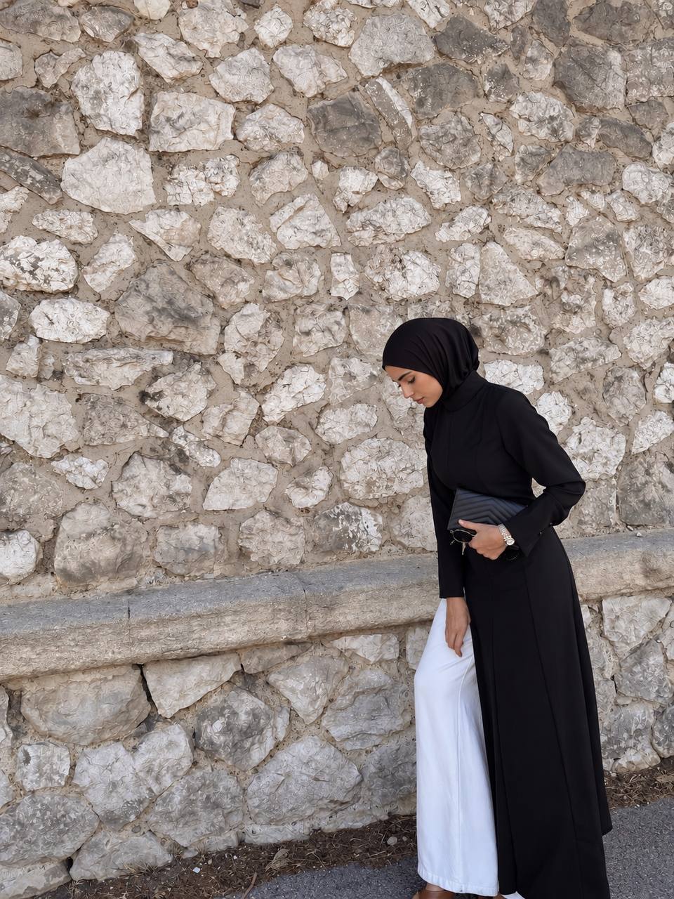 Tunique longue noire L’Aurore portée devant un mur de pierre, tenue femme voilée chic